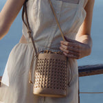 A model wearing a small tan woven vegan leather crossbody bag with a drawstring closure while standing outside.