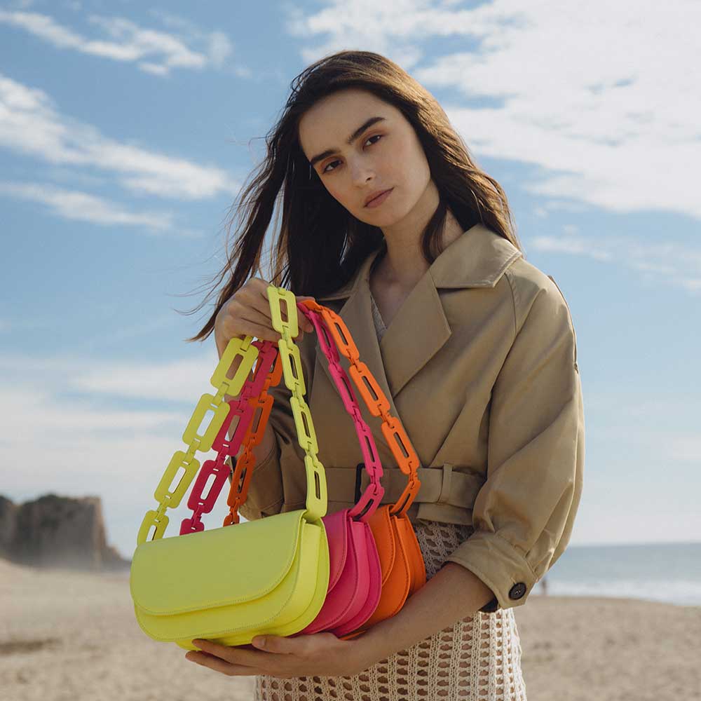 A model wearing three small neon vegan leather shoulder bags with scalloped straps while standing on the beach.
