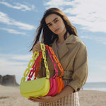A model wearing three neon vegan leather shoulder bags with scalloped straps while standing on the beach