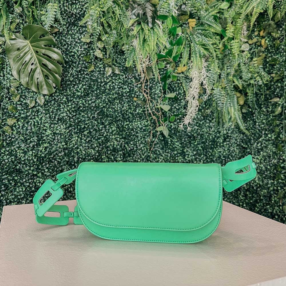 A still image of a neon green vegan leather shoulder bag with a scalloped strap against a greenery background.