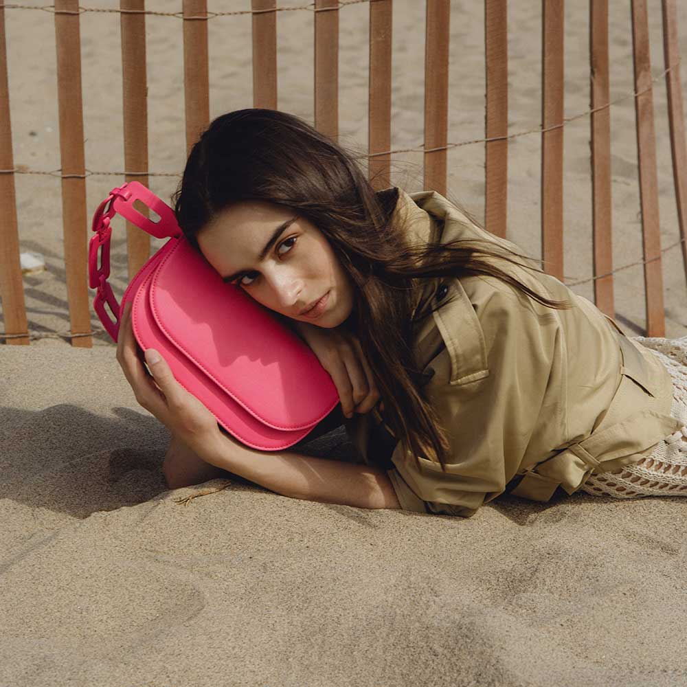 A model wearing a neon pink vegan leather shoulder bag with a scalloped strap while laying in the sand.