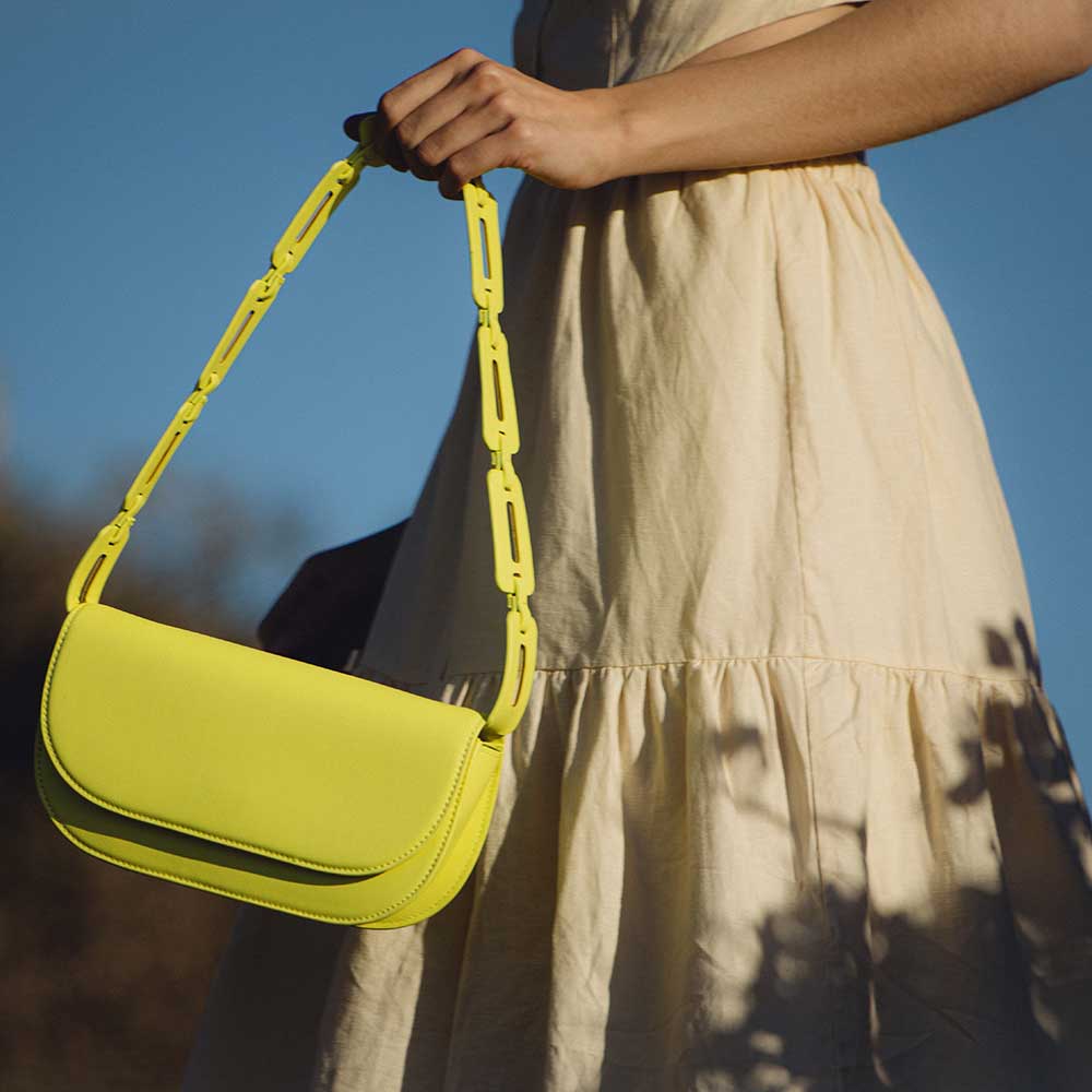 A model wearing a neon yellow vegan leather shoulder bag with a scalloped strap while standing outside.
