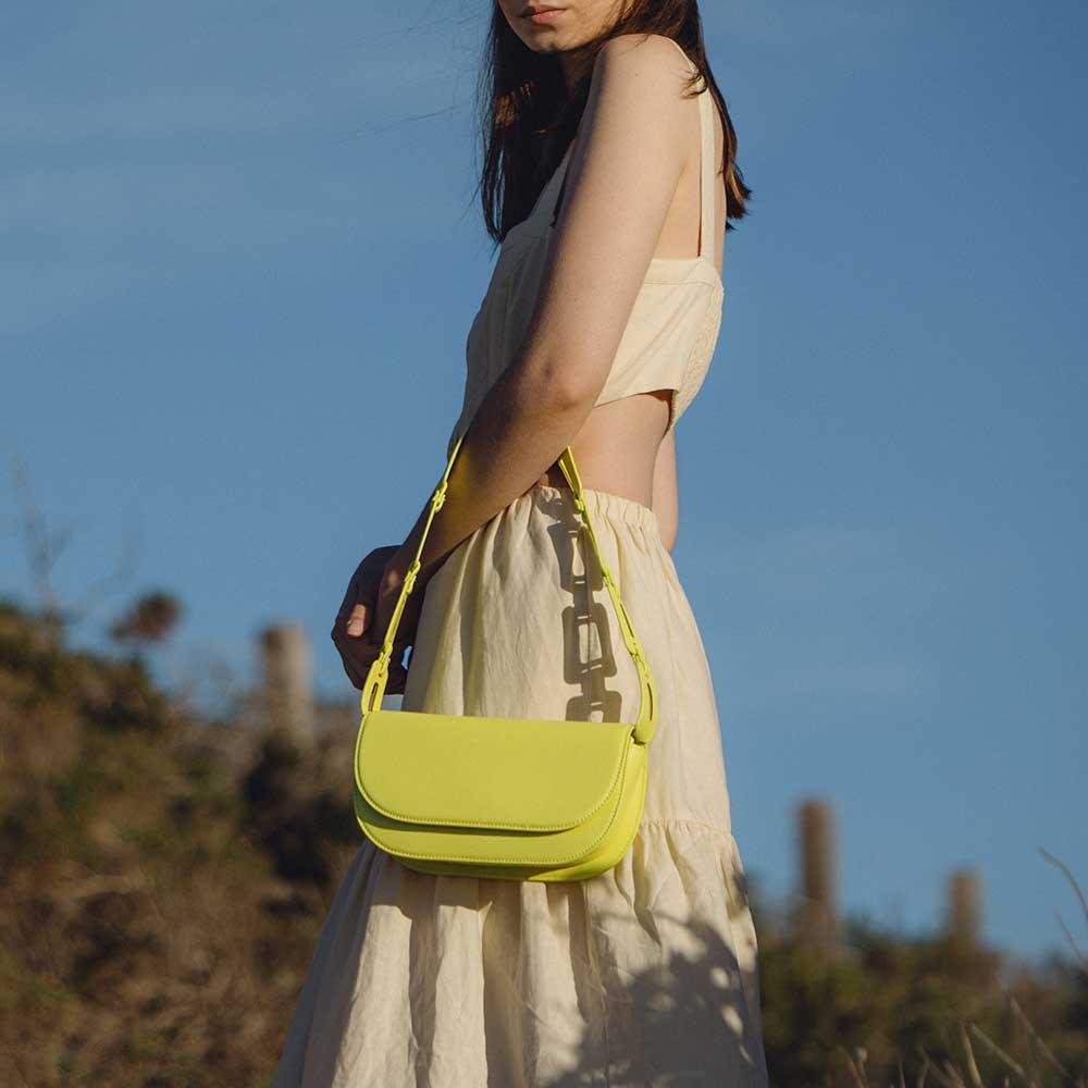 A model wearing a neon yellow vegan leather shoulder bag with a scalloped strap while standing outside.