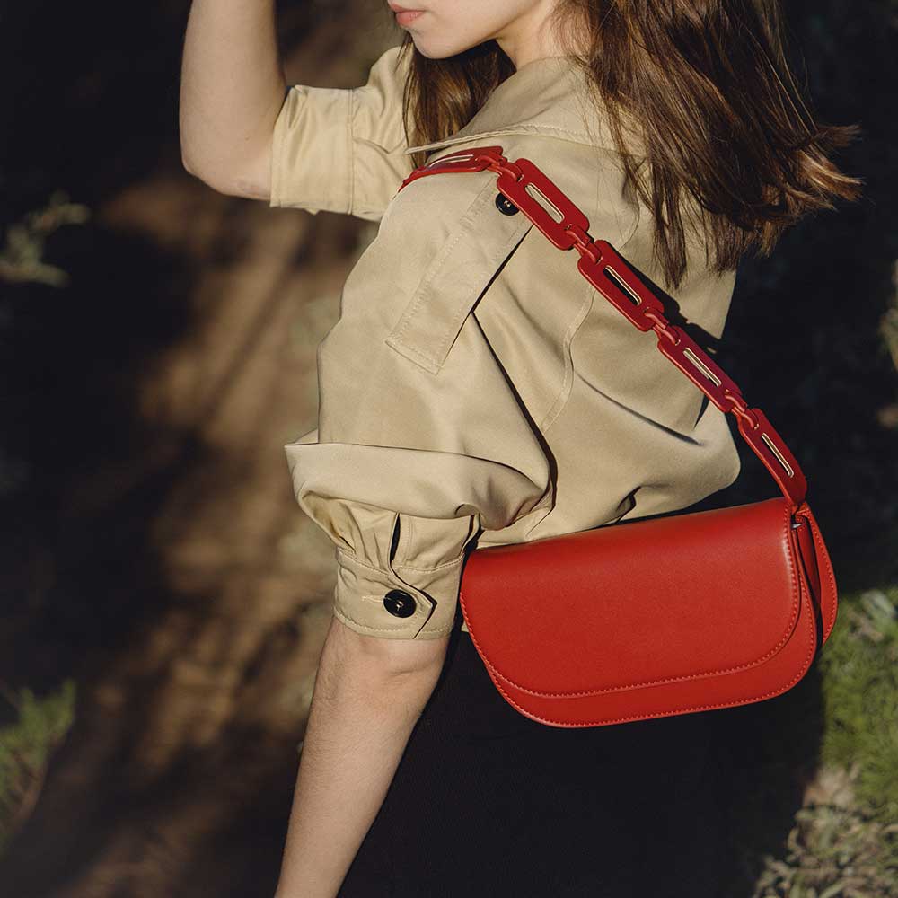 A model wearing a small red vegan leather shoulder bag with a scalloped strap while standing outside.