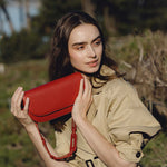 A model wearing a small red vegan leather shoulder bag with a scalloped strap while standing outside.
