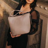 A model holding a taupe recycled vegan leather shoulder bag against a concrete post. 