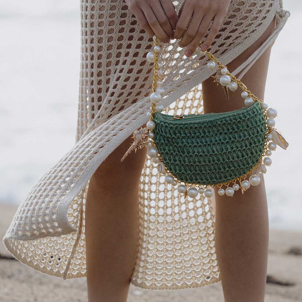 A model wearing a small green crochet straw top handle bag with seashell details along the handle while standing on the beach.