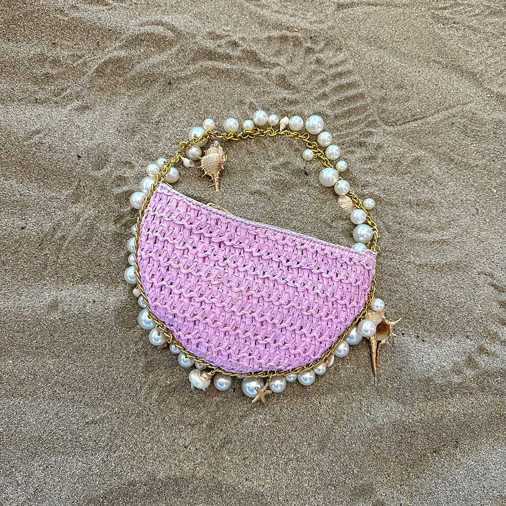 A still image of a small lavender crochet straw top handle bag with seashell details along the handle laying in sand.