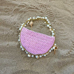 A still image of a small lavender crochet straw top handle bag with seashell details along the handle laying in sand.
