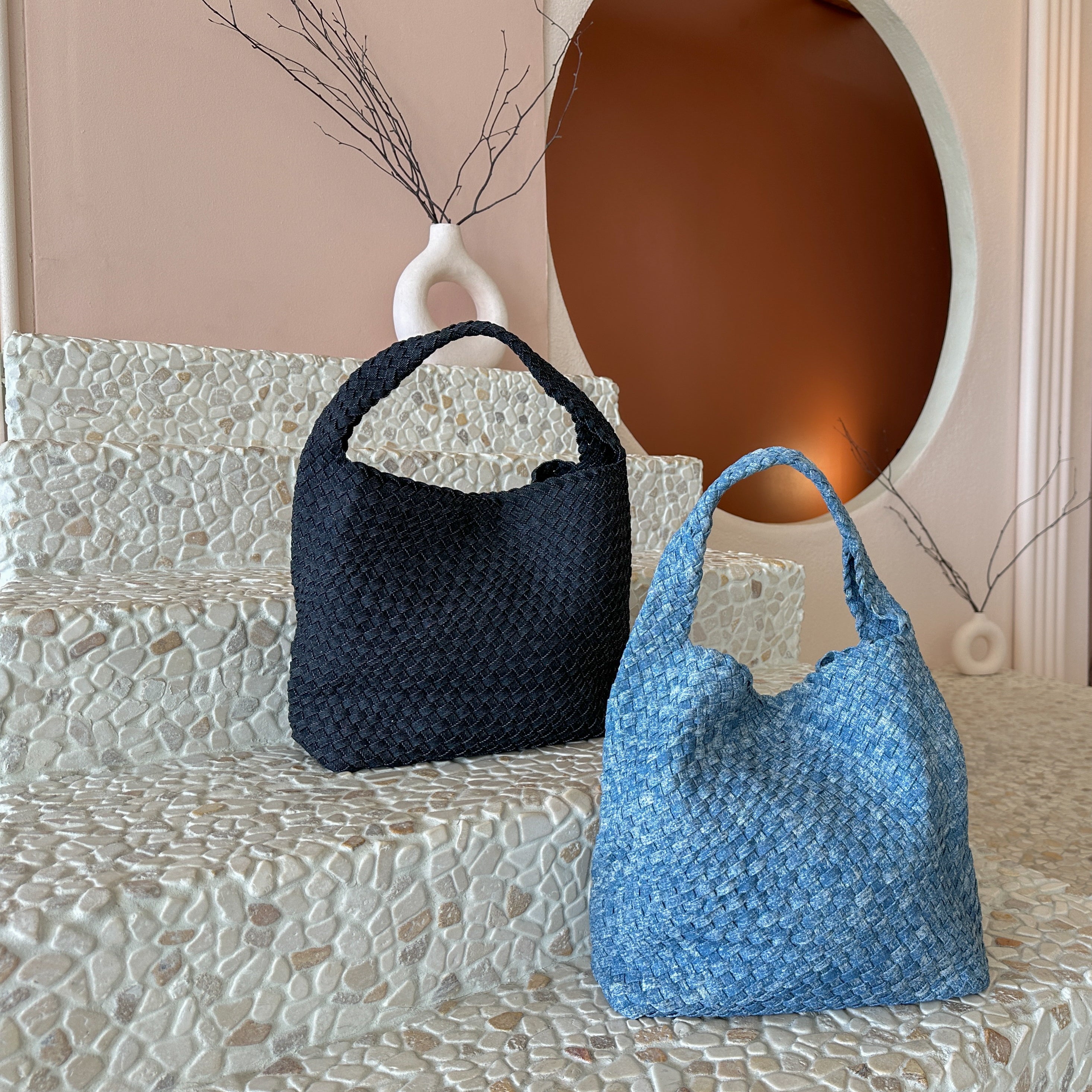 A still image of two denim woven shoulder bags with zip pouch inside laying against a stone background