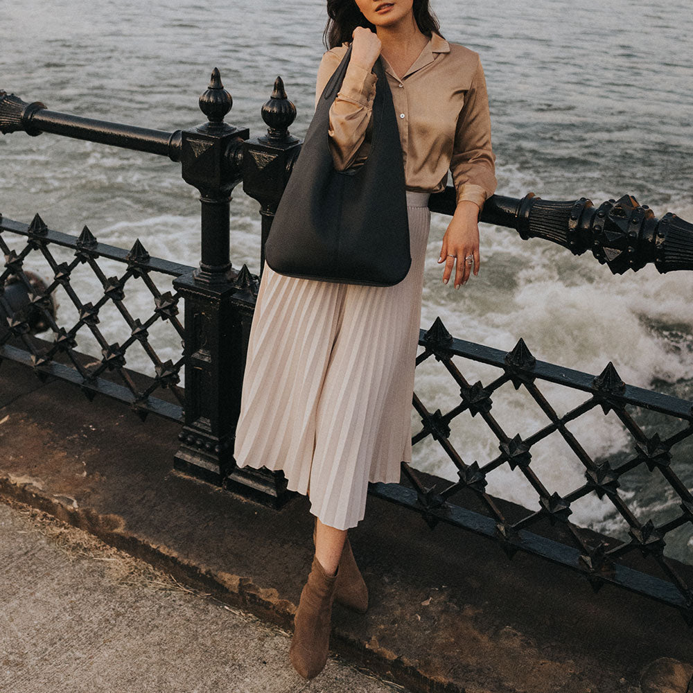 A model wearing a large black recycled vegan leather shoulder bag by the ocean. 