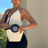 A model wearing a small canvas and vegan leather crossbody bag with a circle handle against a wall outside. 