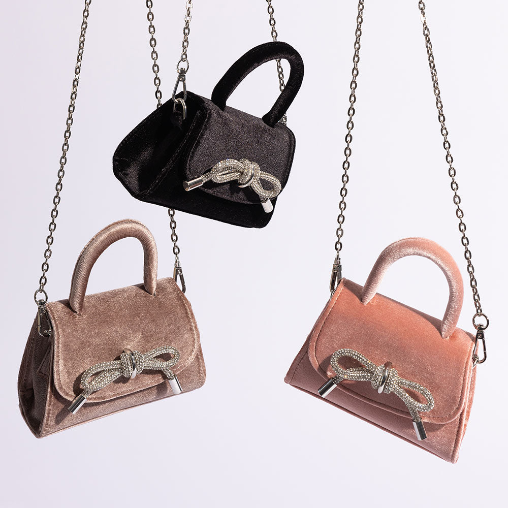 A still image of three mini velvet top handle bags with a silver encrusted bow against a white wall.