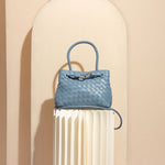 A still image of a sky hand woven vegan leather crossbody bag against a tan wall. 