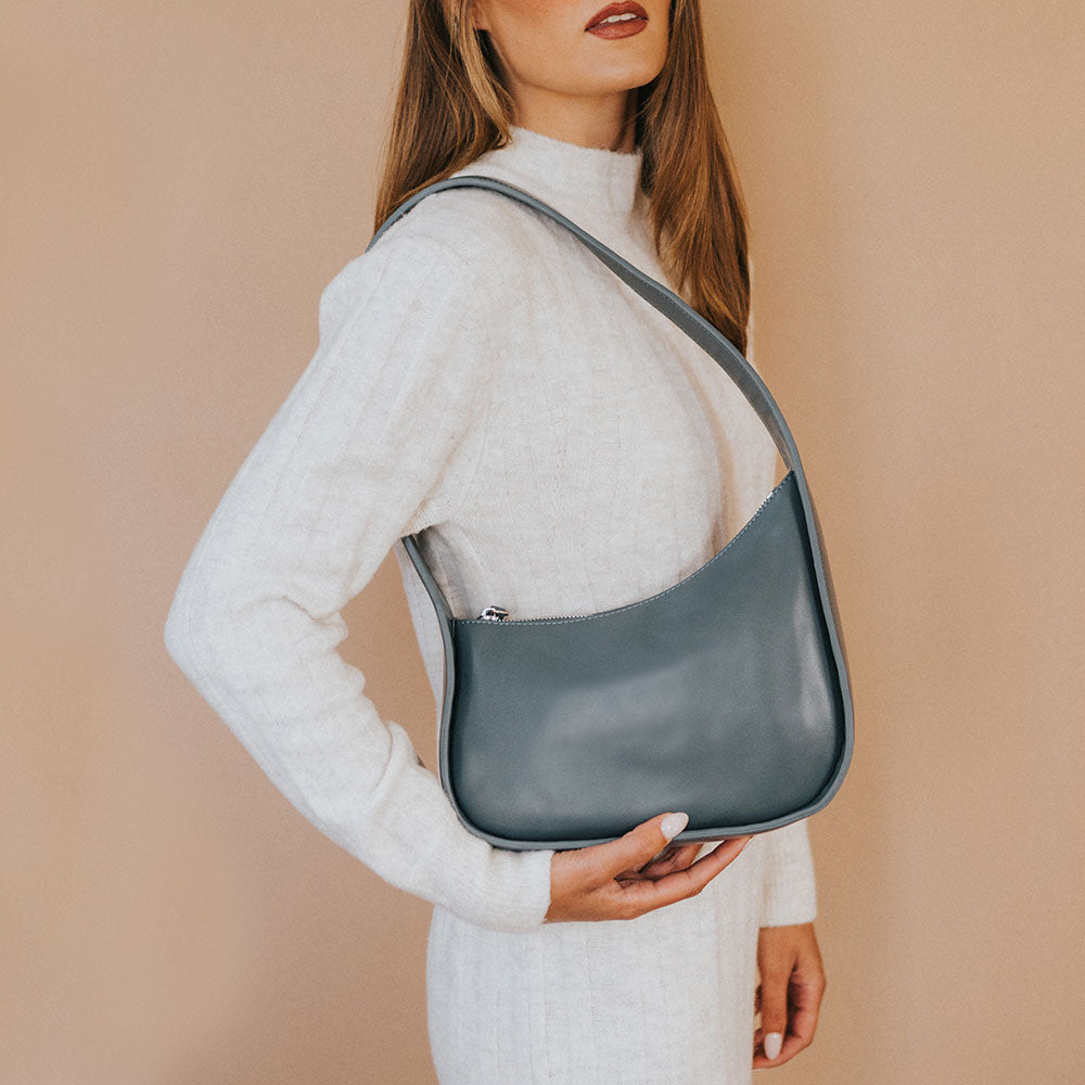 A model wearing a asymmetrical vegan leather structured shoulder bag against a tan wall. 