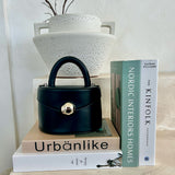 A still image of a small recycled vegan leather top handle bag with silver hardware with books and a vase.