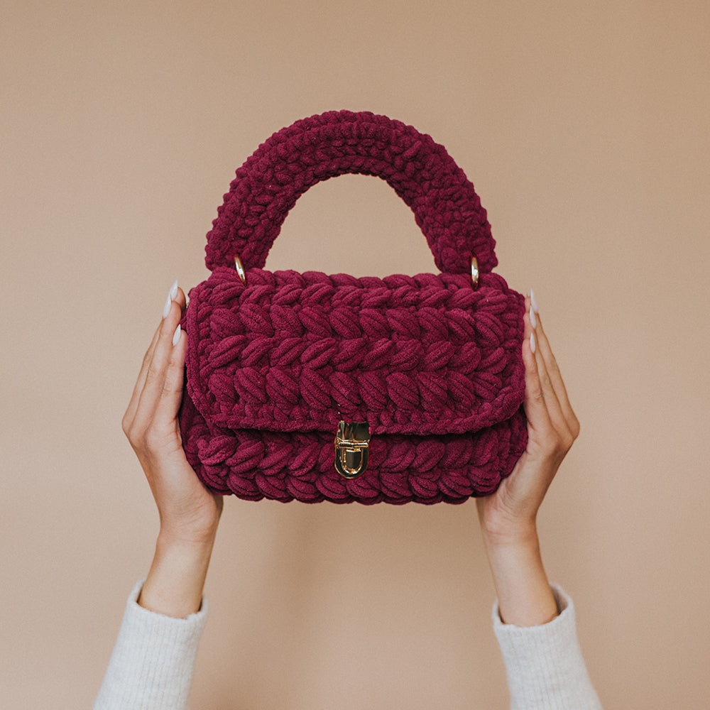 A model holding up a plum knitted handbag with a gold clasp against a blank wall. 