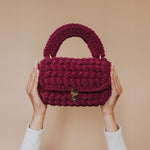 A model holding up a plum knitted handbag with a gold clasp against a blank wall. 