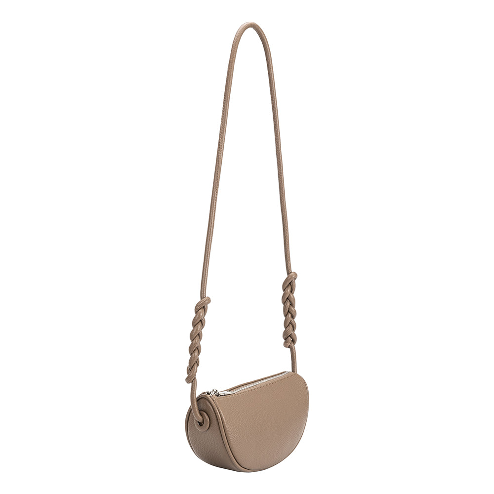 A small taupe crescent shaped vegan leather crossbody bag with a braided handle.