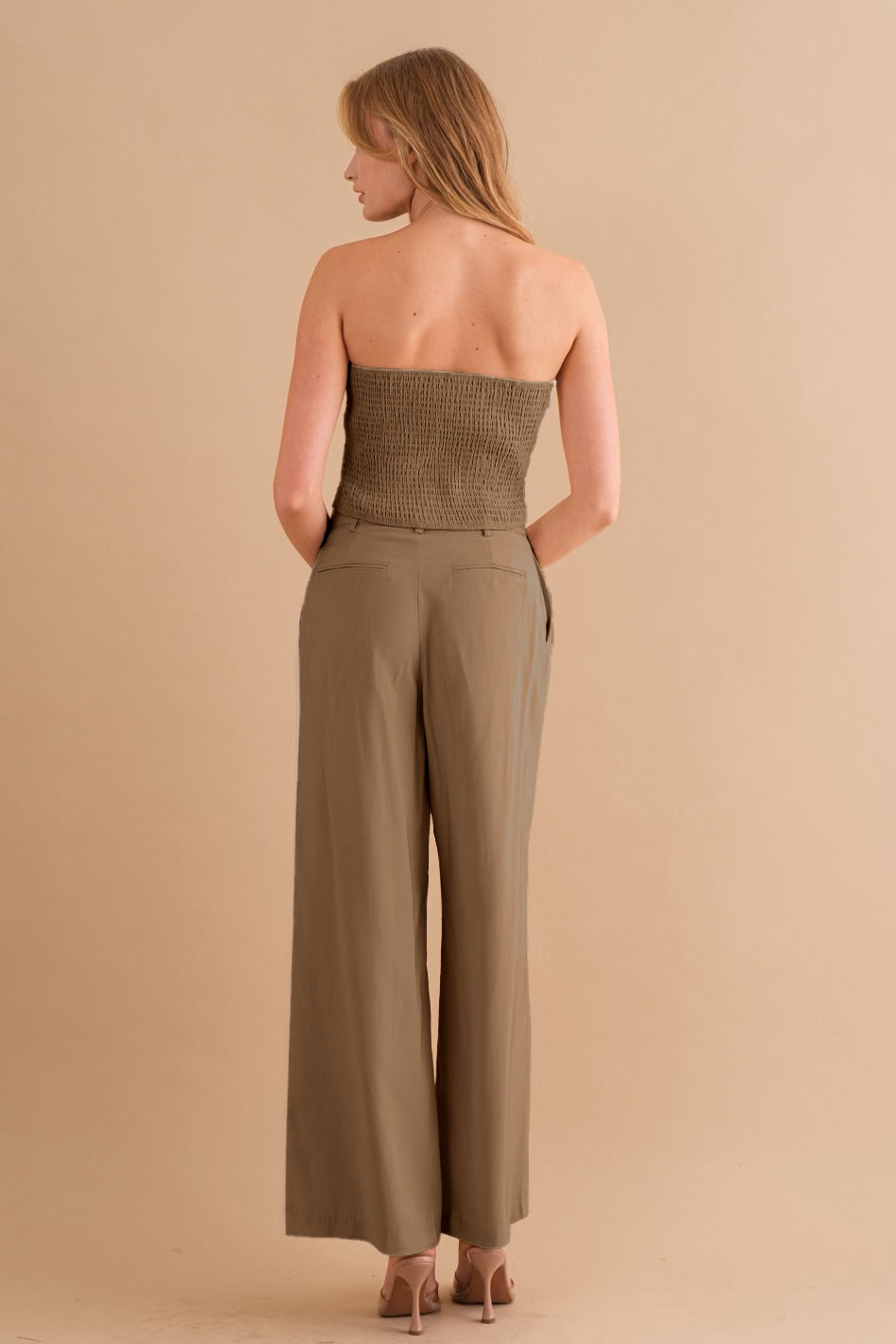 A backside view of a model wearing a two piece strapless top and pant set against a tan wall. 