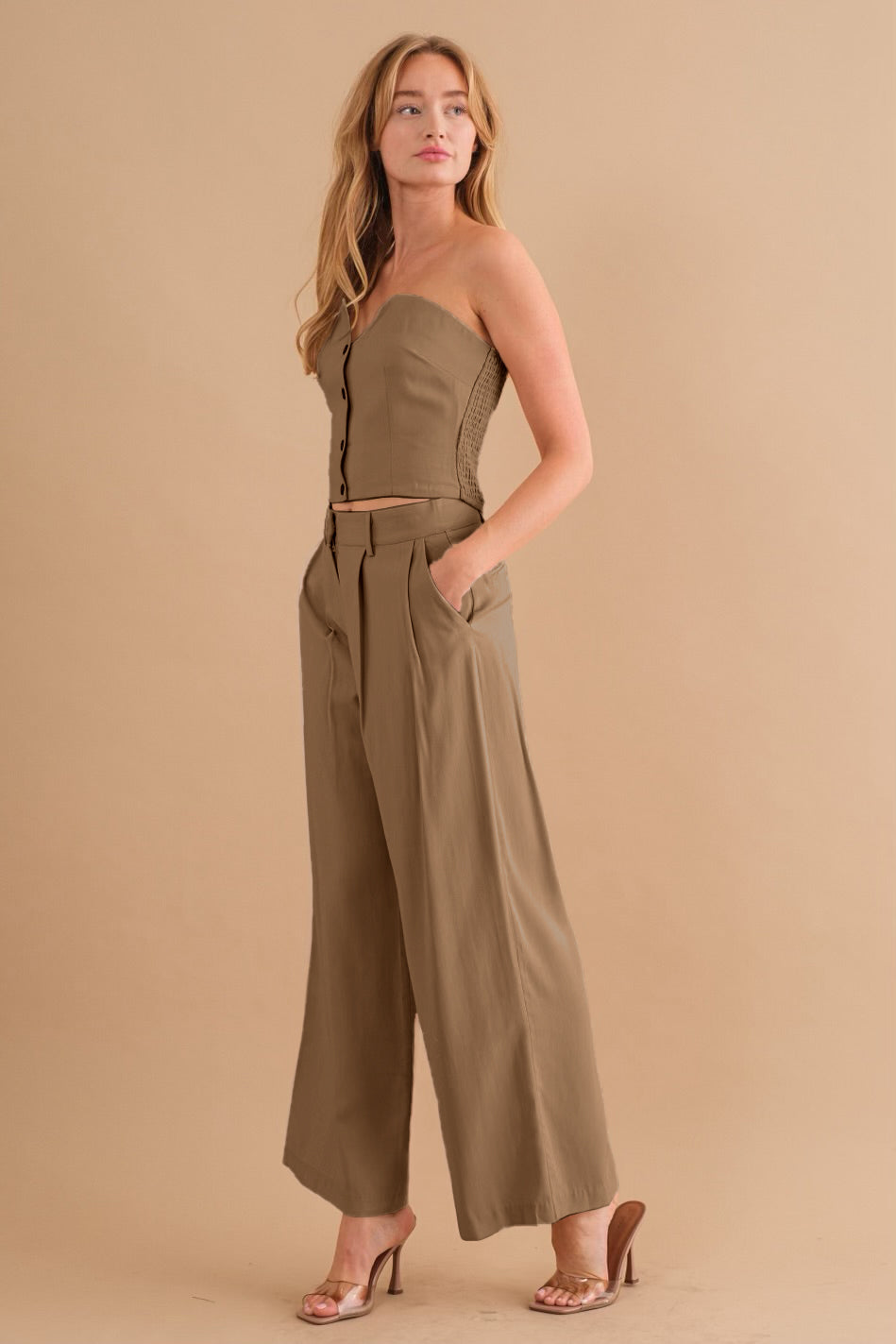A model wearing a tan two piece strapless top and pant set against a tan wall. 