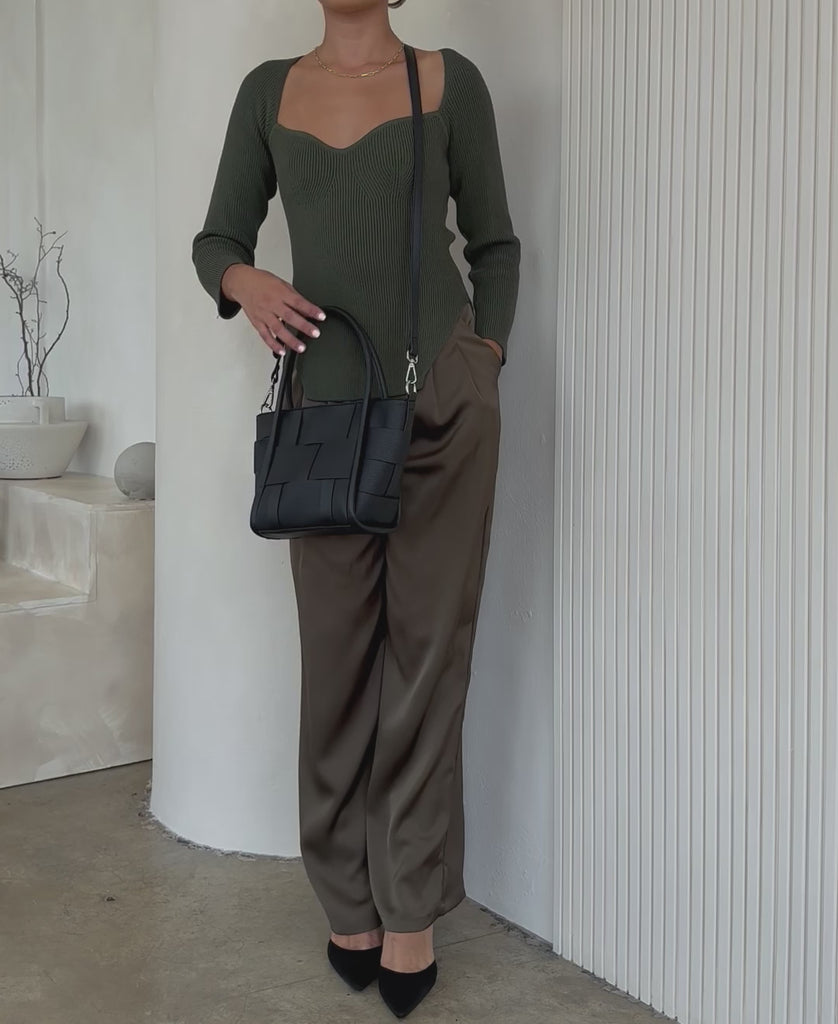 Video of a model wearing a small woven wide strap tote bag against a white wall. 