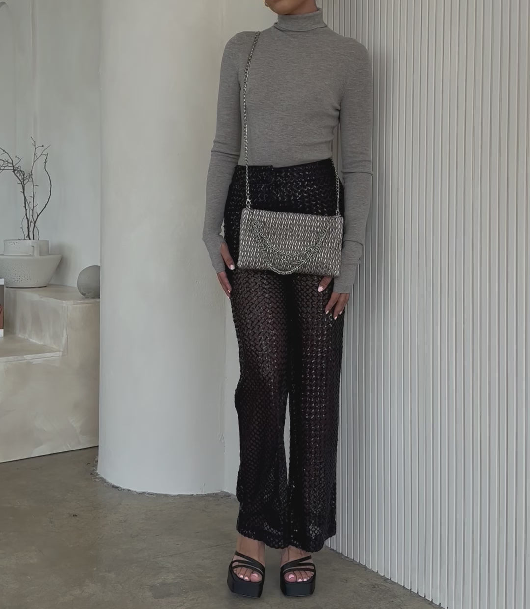 Video of a model wearing a small quilted pattern clutch with silver chain against a white wall. 