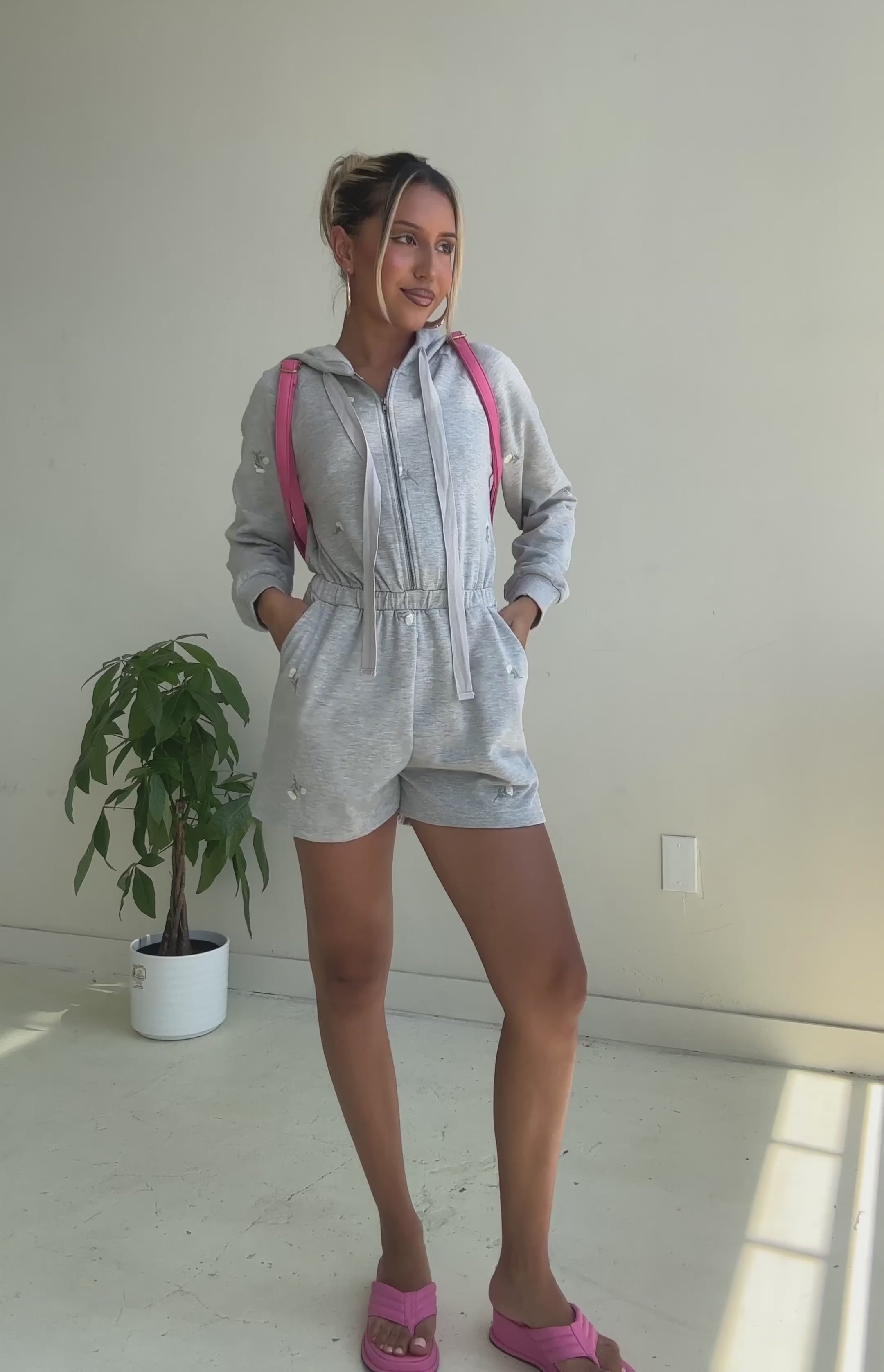 Video fo a model wearing a grey terry hooded romper against a white wall. 