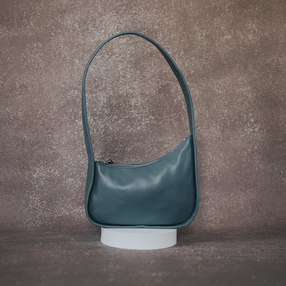 A still image of a asymmetrical vegan leather structured shoulder bag against a grey wall. 