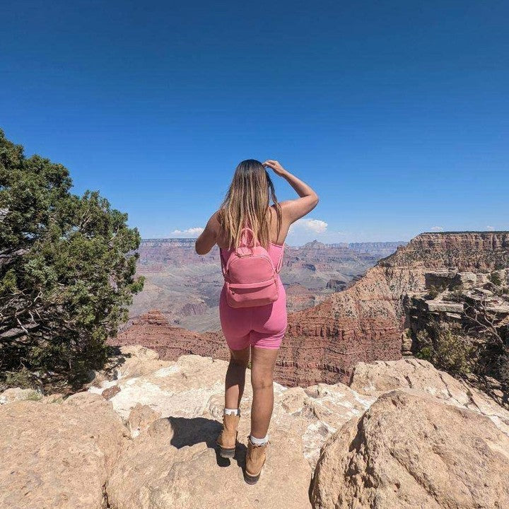 A model wearing a pink recycled vegan leather backpack with a front zip pocket while outside standing on rocks.