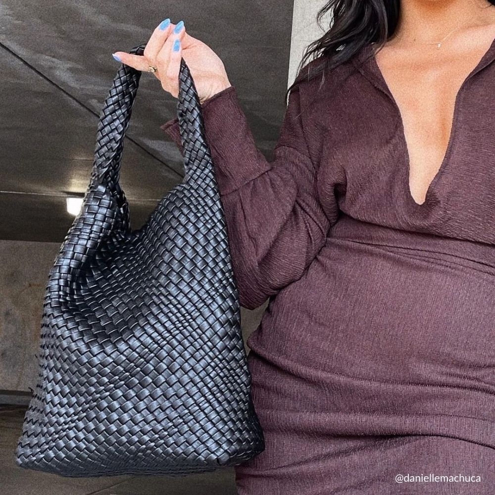A model holding a large black woven vegan leather shoulder bag outside in a parking garage. 