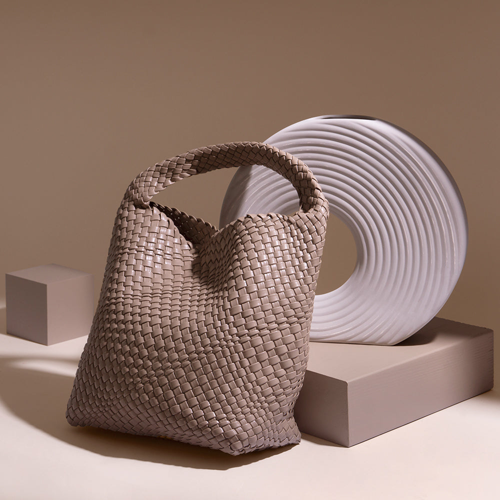 A still image of a large woven vegan leather shoulder bag against a tan wall with a white vase. 