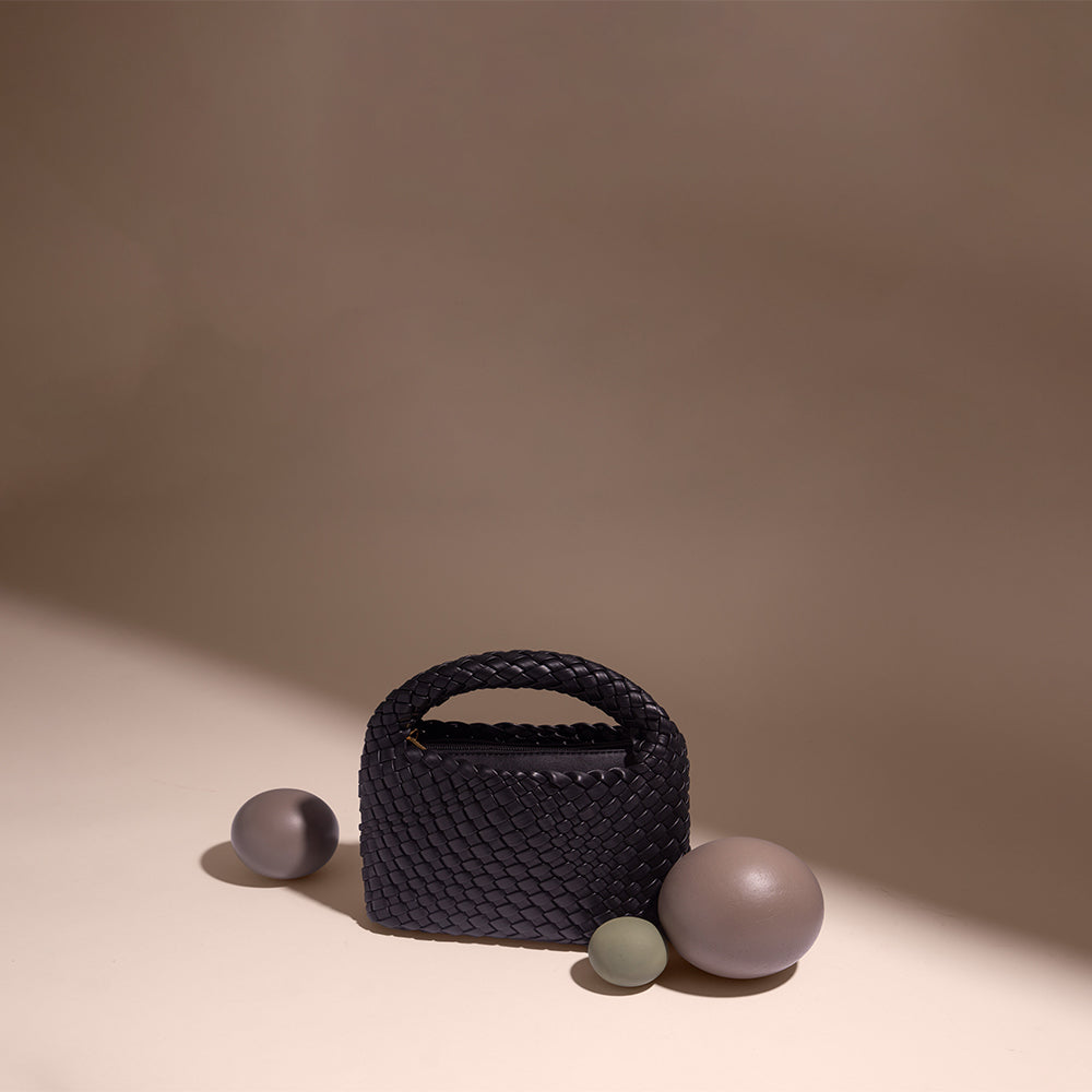 A still image of a small handwoven vegan leather top handle bag against a brown wall. 