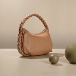 A still image of a small tan recycled vegan leather shoulder bag with a braided handle against a tan background with green ball props. 