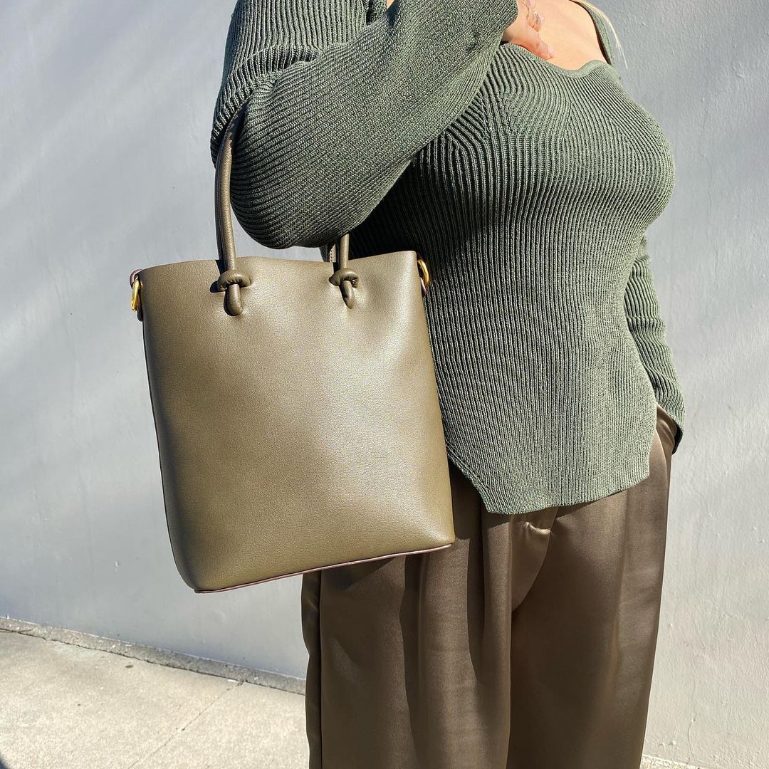 A model wearing a sweetheart rib knit long sleeve top against a white wall with a vegan leather handbag.