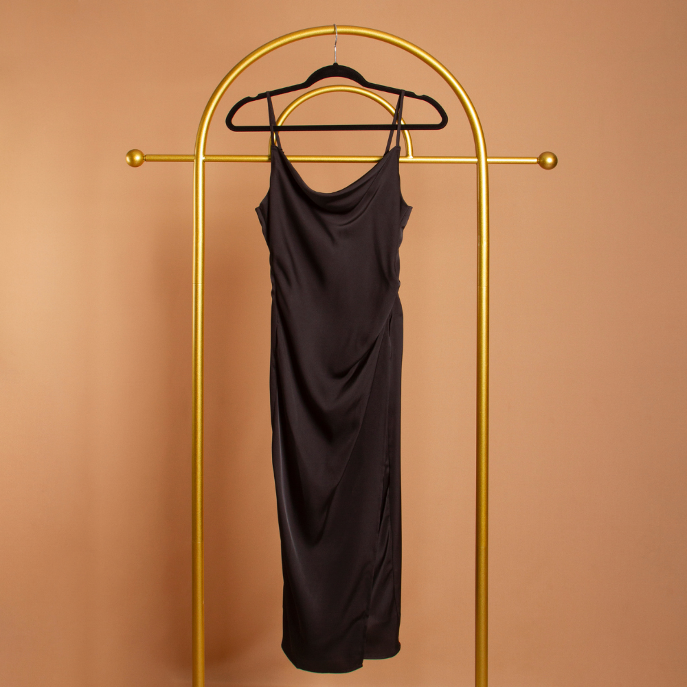 A black satin cowl neck slip dress on a hanger with an orange background. 