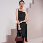 A model wearing a black satin cowl dress while holding a burgundy shoulder bag with a beige background. 