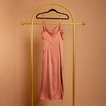 A ginger satin cowl neck slip dress backside view on a hanger with a orange background. 