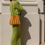 A model wearing a green two piece cardigan and midi dress set while holding a pleated shoulder bag against a white wall.