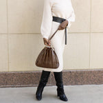 Model wearing a chocolate pleated vegan leather shoulder bag against a stone wall. 