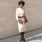 Model wearing a chocolate pleated vegan leather shoulder bag. Holding a chocolate zip ouch against a cream wall.