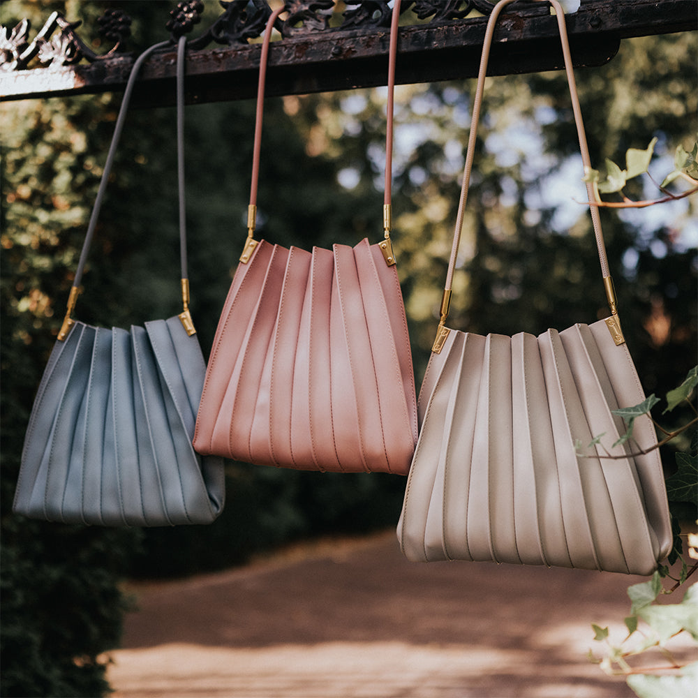 Still photo of three pleated vegan leather shoulder bags hanging on a metal fence outside. 