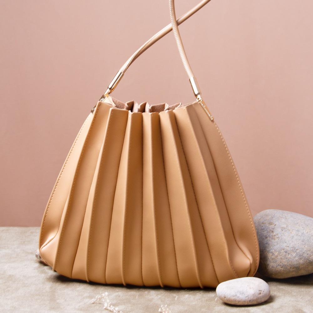 A still image of a taupe pleated vegan leather shoulder bag with stone decorations against a pink wall. 