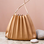 A still image of a taupe pleated vegan leather shoulder bag with stone decorations against a pink wall. 