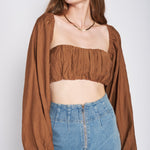 A model wearing a woven tan crop top and shrug set against a white wall. 