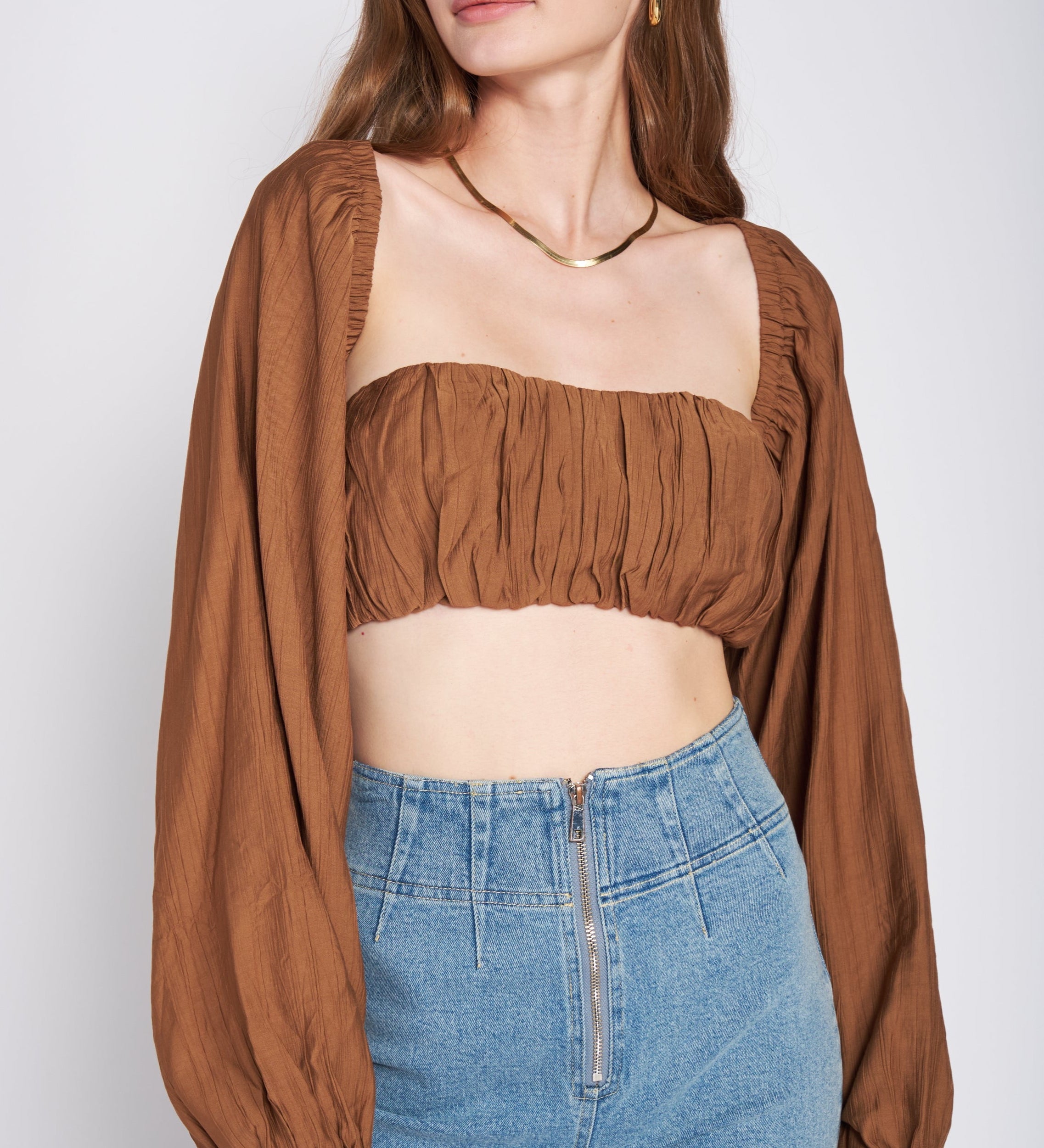 A model wearing a woven tan crop top and shrug set against a white wall. 