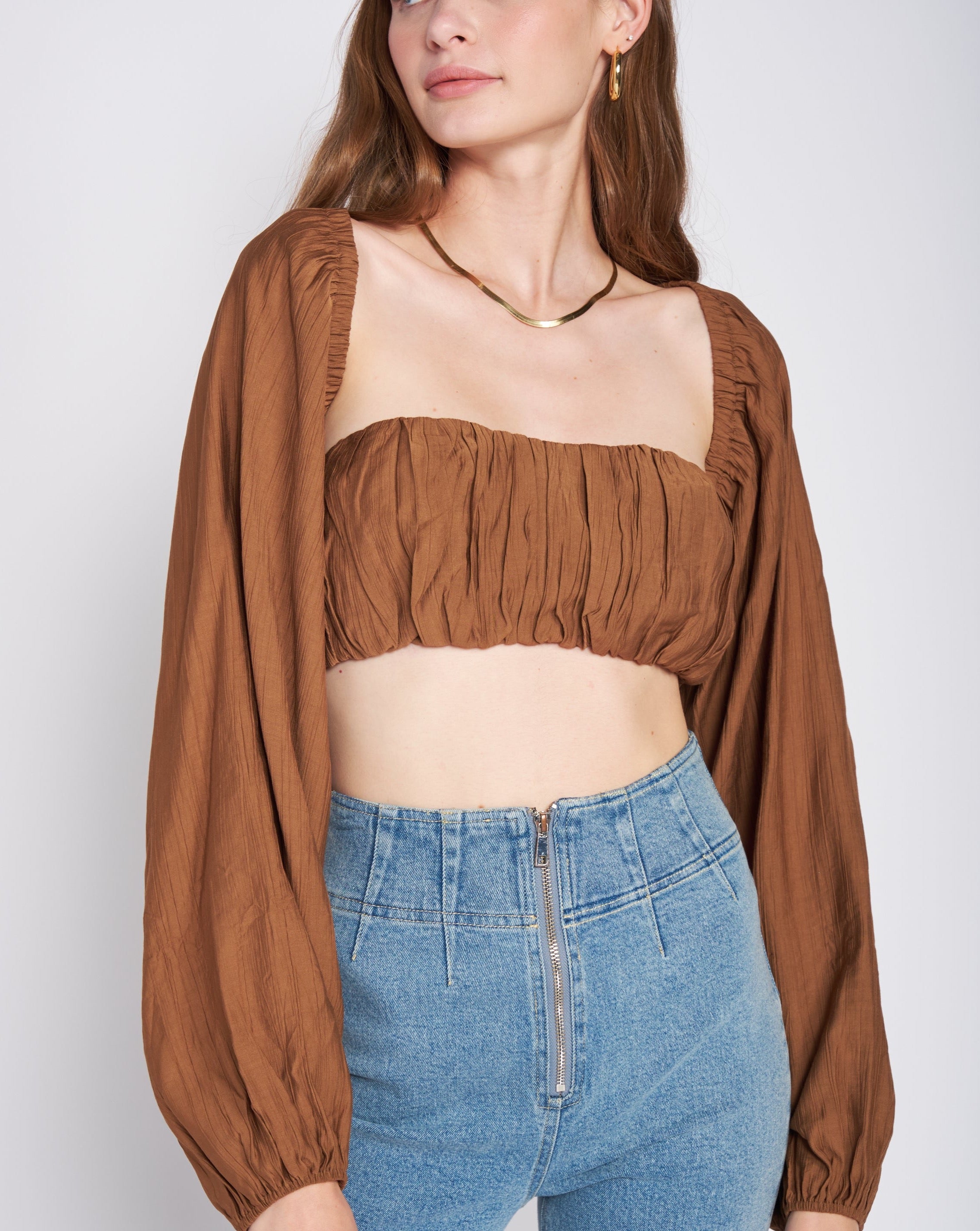 A model wearing a woven tan crop top and shrug set against a white wall. 