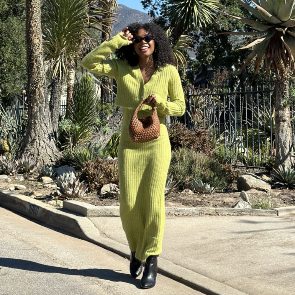 A model wearing a two piece green cardigan and midi dress while holding a woven bag outside. 