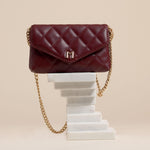 A still image of a small burgundy quilted vegan leather shoulder bag against a white wall. 