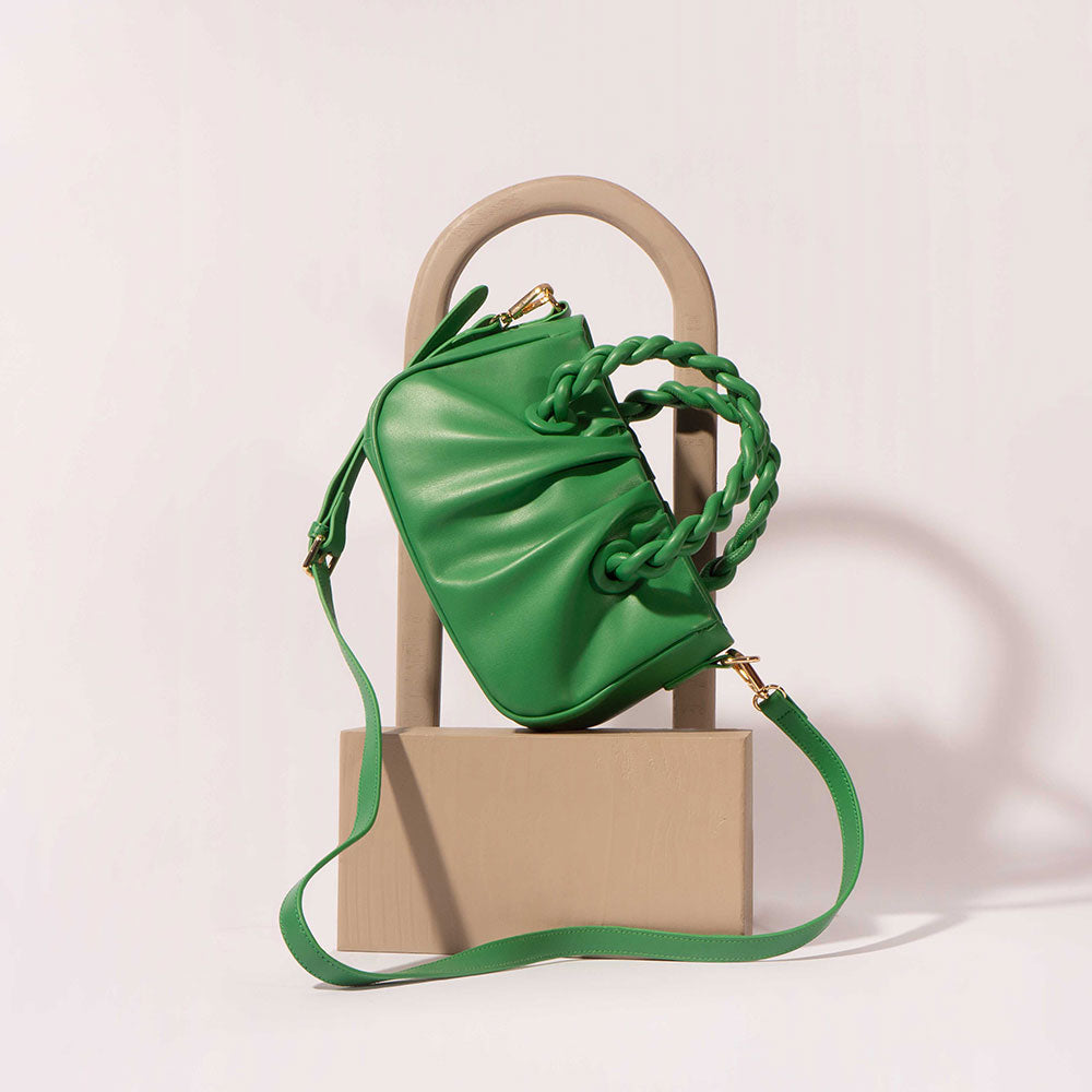 A small green recycled vegan leather crossbody bag with a twisted strap against a white wall with tan props. 